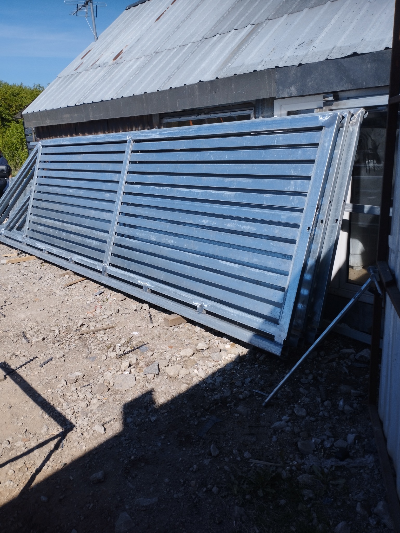 Szara, metalowa brama wjazdowa z poziomymi szczeblami, oparta o budynek z blachy falistej. Widoczne ślady spawania. Brama leży na żwirowym podłożu.