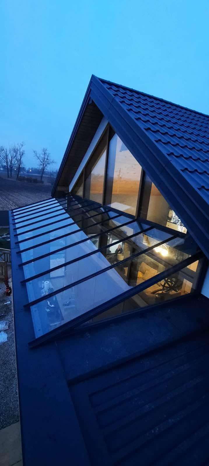 Szklana zabudowa balkonu z czarną ramą, widok z góry na konstrukcję i wnętrze. W tle pola i drzewa o zmierzchu. Nietypowy kształt dachu.