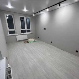 IOMA GROUP SPÓŁKA Z OGRANICZONĄ ODPOWIEDZIALNOŚCIĄ - Jasne, szare wnętrze po remoncie: panele podłogowe, gładkie ściany, okna z grzejnikami. Minimalistyczne oświetlenie sufitowe.