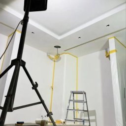 IOMA GROUP SPÓŁKA Z OGRANICZONĄ ODPOWIEDZIALNOŚCIĄ - Białe ściany pokoju zabezpieczone taśmą malarską i folią, przygotowane do malowania. Widoczna drabina, lampa na statywie i akcesoria malarskie.
