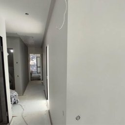 IOMA GROUP SPÓŁKA Z OGRANICZONĄ ODPOWIEDZIALNOŚCIĄ - Jasne, świeżo pomalowane ściany korytarza w odcieniach szarości i bieli. Widoczne gniazdko elektryczne i przewody. W tle otwarte drzwi i okno.