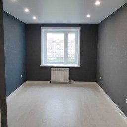 IOMA GROUP SPÓŁKA Z OGRANICZONĄ ODPOWIEDZIALNOŚCIĄ - Pomalowane na szaro wnętrze pokoju z białymi panelami podłogowymi i grzejnikiem pod oknem. Sufit z oświetleniem punktowym. Widok na budynki za oknem.