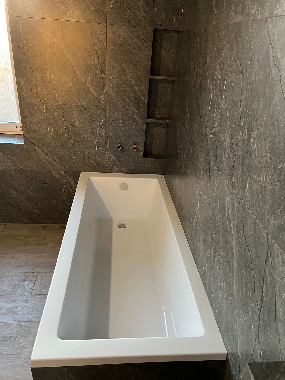 Nowoczesna, biała wanna w łazience wykończonej szarą, marmurkową płytką. W ścianie wnęka z półkami. Widoczne przyłącza wodne. Minimalistyczny design.