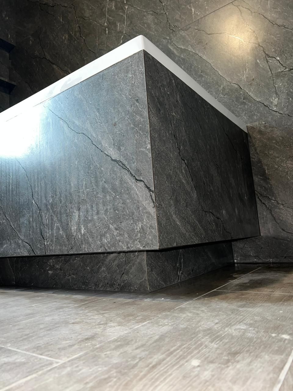 Obudowana, nowoczesna wanna z szarego kamienia z białym wykończeniem, dopasowana do płytek na ścianach i podłodze. Minimalistyczny design łazienki.