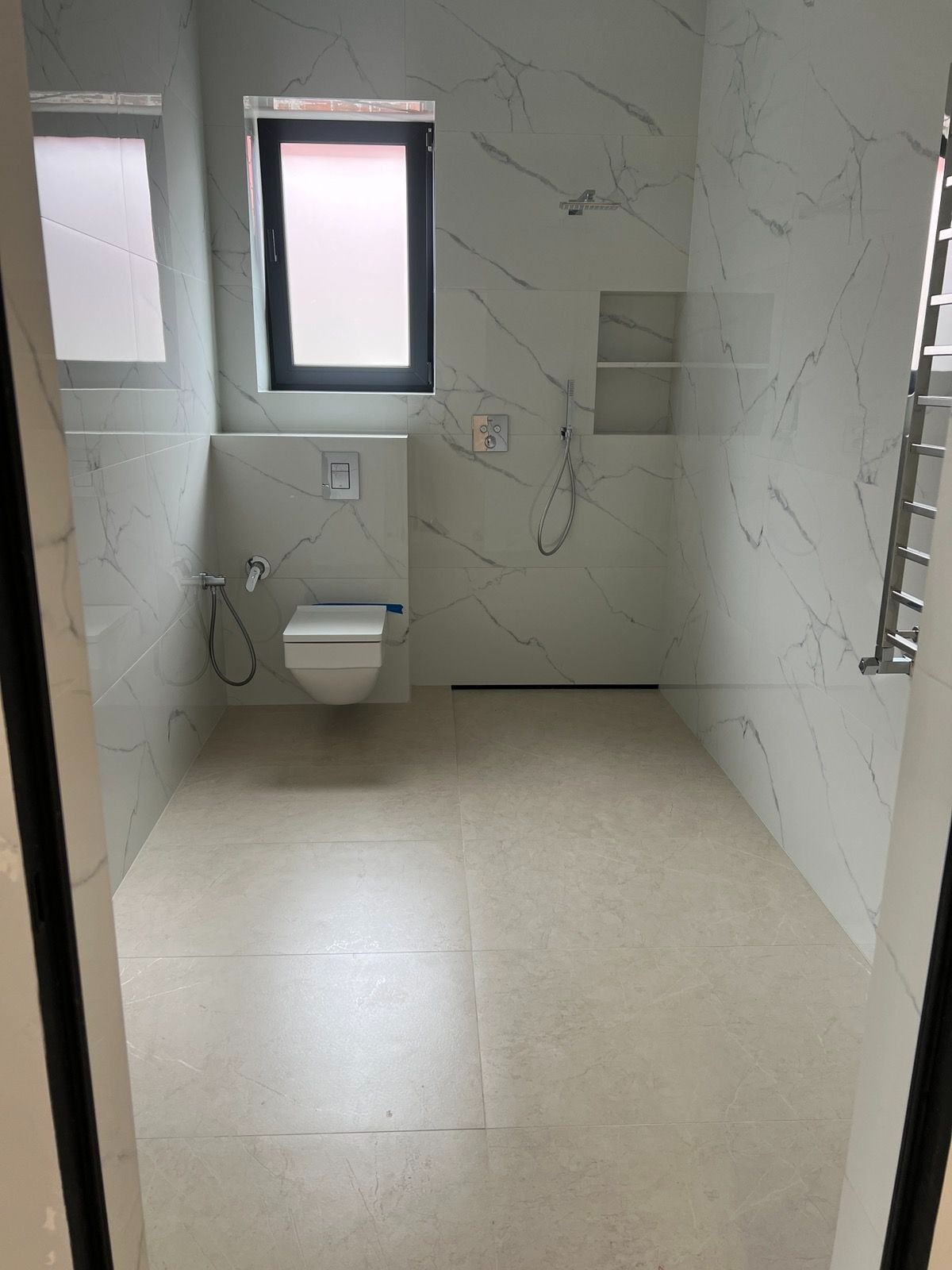 Nowoczesna łazienka z białymi płytkami imitującymi marmur, beżowa podłoga, czarna rama okna. Minimalistyczny design, widok zza otwartych drzwi.