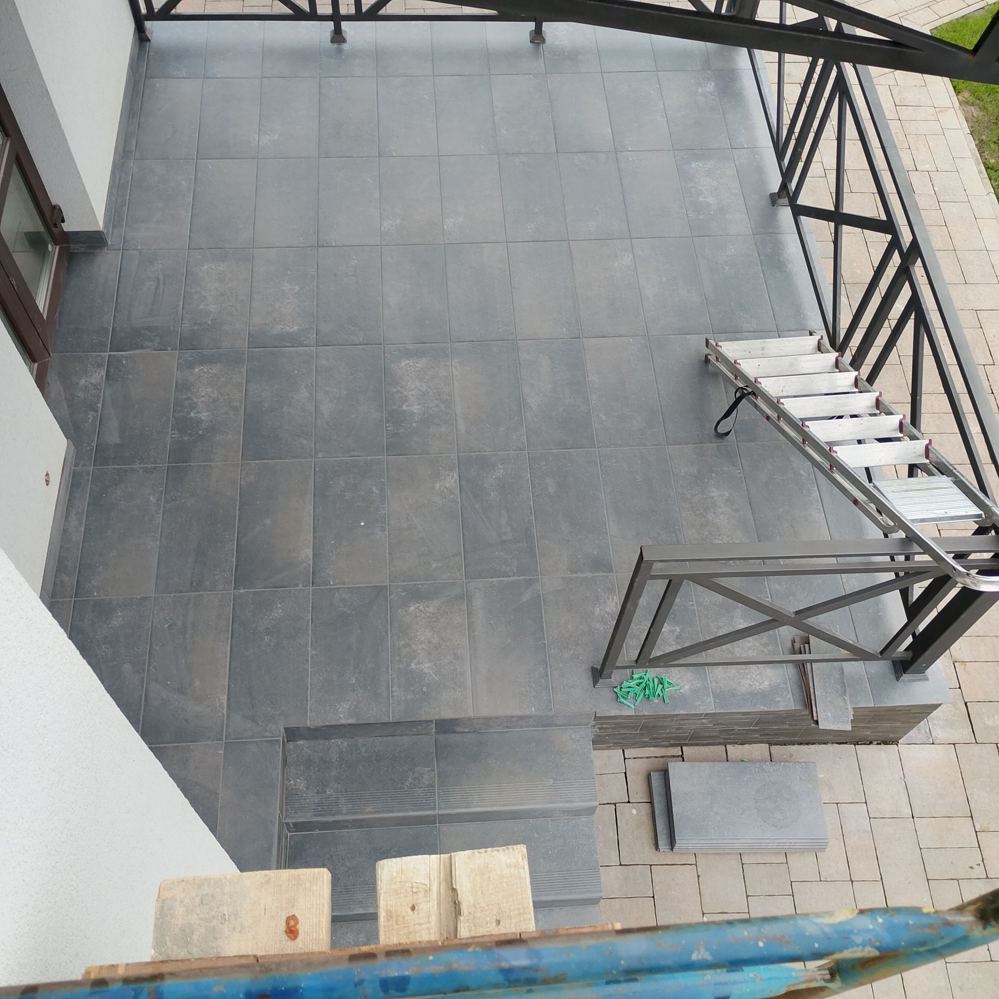 Wykończony taras z szarych płytek, widok z góry. Metalowa konstrukcja z barierkami, drabina i zapas płytek. Detale montażowe widoczne obok schodów.