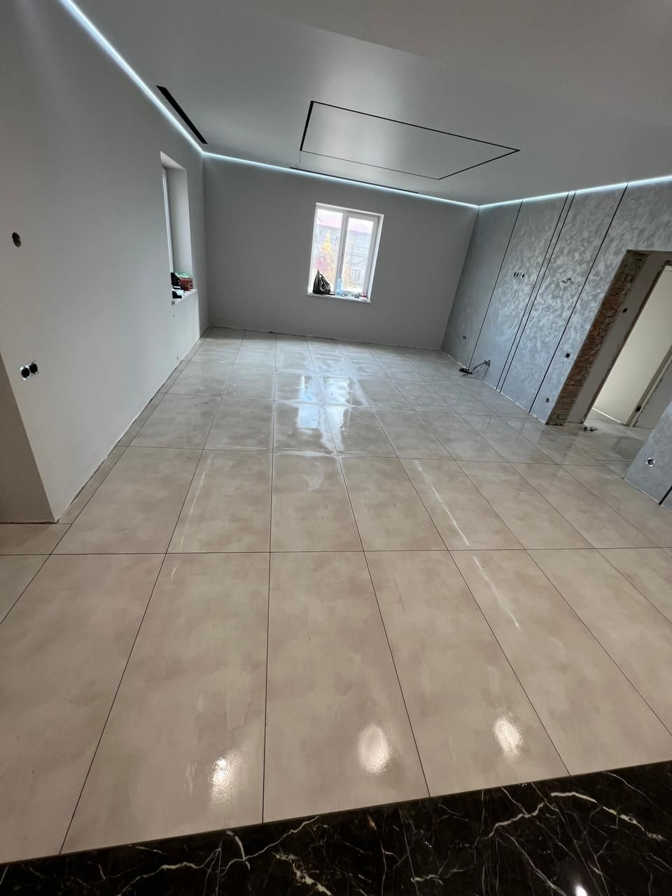 Wykończone wnętrze z beżowymi płytkami podłogowymi i dekoracyjną ścianą. Widoczne oświetlenie LED podsufitowe i okno z parapetem. Drzwi w stanie surowym.