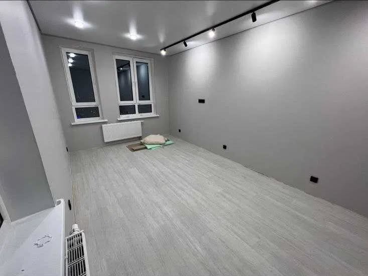 Jasne, szare wnętrze po remoncie: panele podłogowe, gładkie ściany, okna z grzejnikami. Minimalistyczne oświetlenie sufitowe.