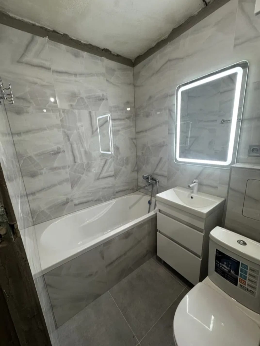 Wykończona łazienka z wanną, umywalką z szafką i toaletą. Na ścianach szare płytki imitujące marmur, podświetlane lustra. Widoczne niedociągnięcia na suficie.