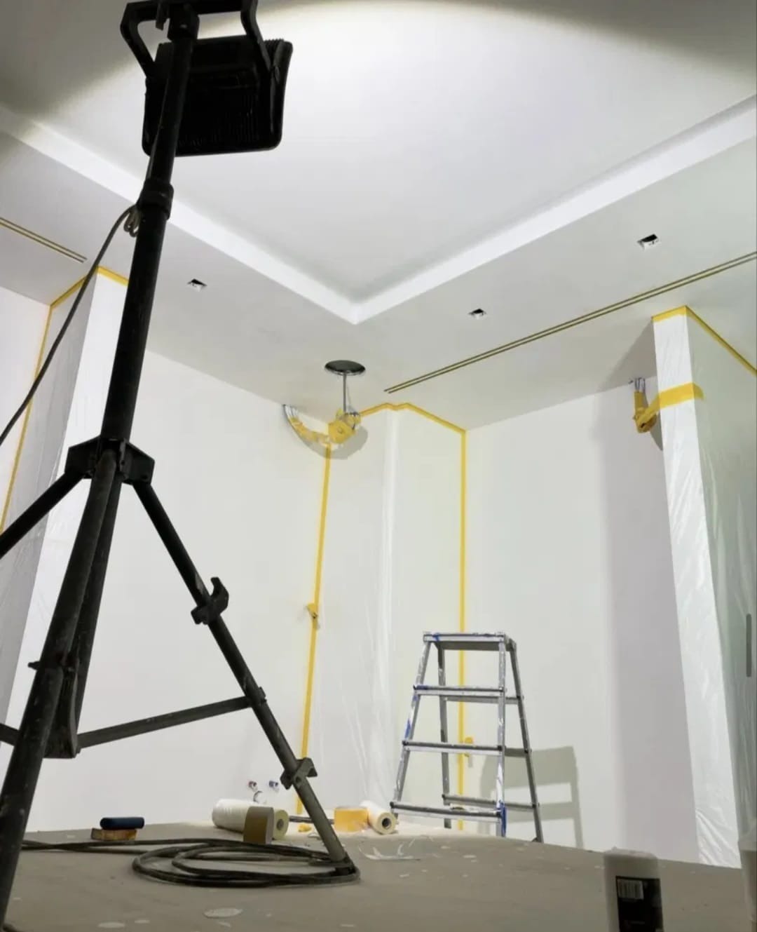 Białe ściany pokoju zabezpieczone taśmą malarską i folią, przygotowane do malowania. Widoczna drabina, lampa na statywie i akcesoria malarskie.
