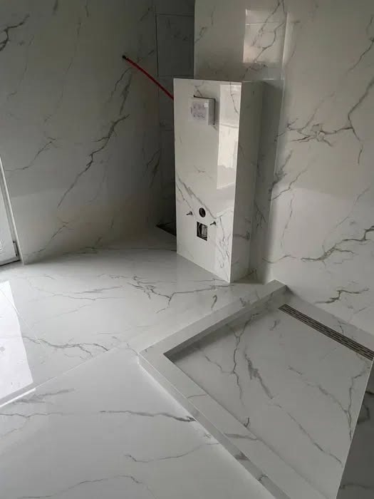 Biała łazienka z marmurowymi płytkami, widoczny brodzik i zabudowa WC. Minimalistyczny design, błyszczące powierzchnie, nowoczesne wykończenie.