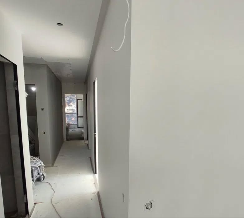 Jasne, świeżo pomalowane ściany korytarza w odcieniach szarości i bieli. Widoczne gniazdko elektryczne i przewody. W tle otwarte drzwi i okno.