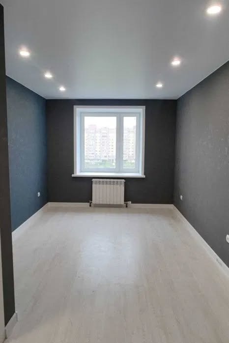 Pomalowane na szaro wnętrze pokoju z białymi panelami podłogowymi i grzejnikiem pod oknem. Sufit z oświetleniem punktowym. Widok na budynki za oknem.