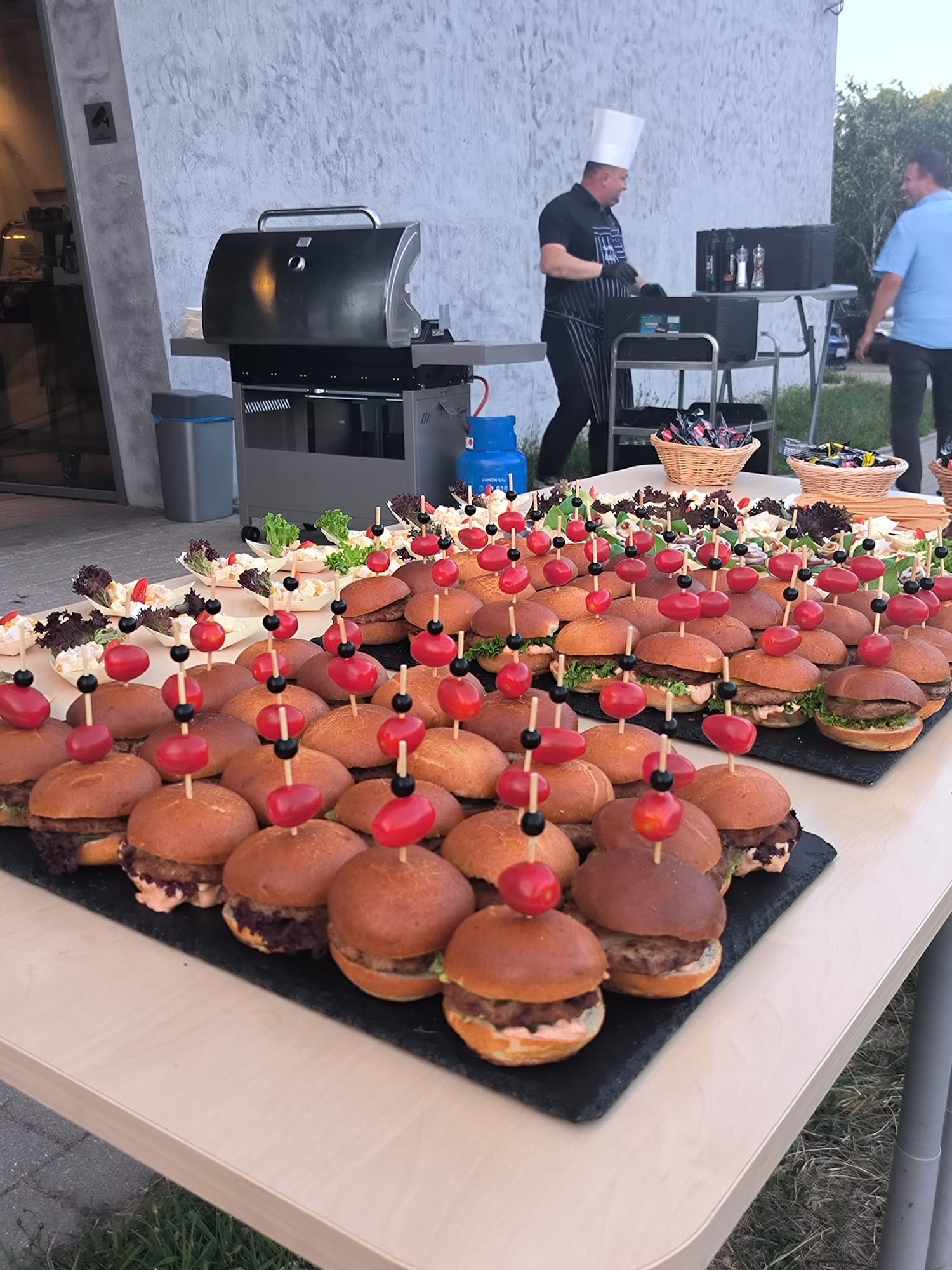 Stół z mini burgerami i przekąskami, przygotowany przez kucharza w tle, grill obok. Catering na imprezę plenerową z dbałością o detale.
