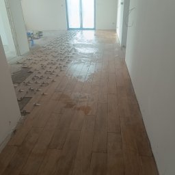 Krzysztof Matusiak KrisBud - Układanie podłogi z płytek imitujących drewno w jasnym wnętrzu. Widoczne klipsy poziomujące i fragmenty nieukończonej podłogi. W tle okno balkonowe.