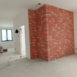 Krzysztof Matusiak KrisBud - Ściana wykończona czerwoną cegłą klinkierową w rogu pomieszczenia. Widoczne surowe ściany i podłoga, sugerujące etap prac wykończeniowych.