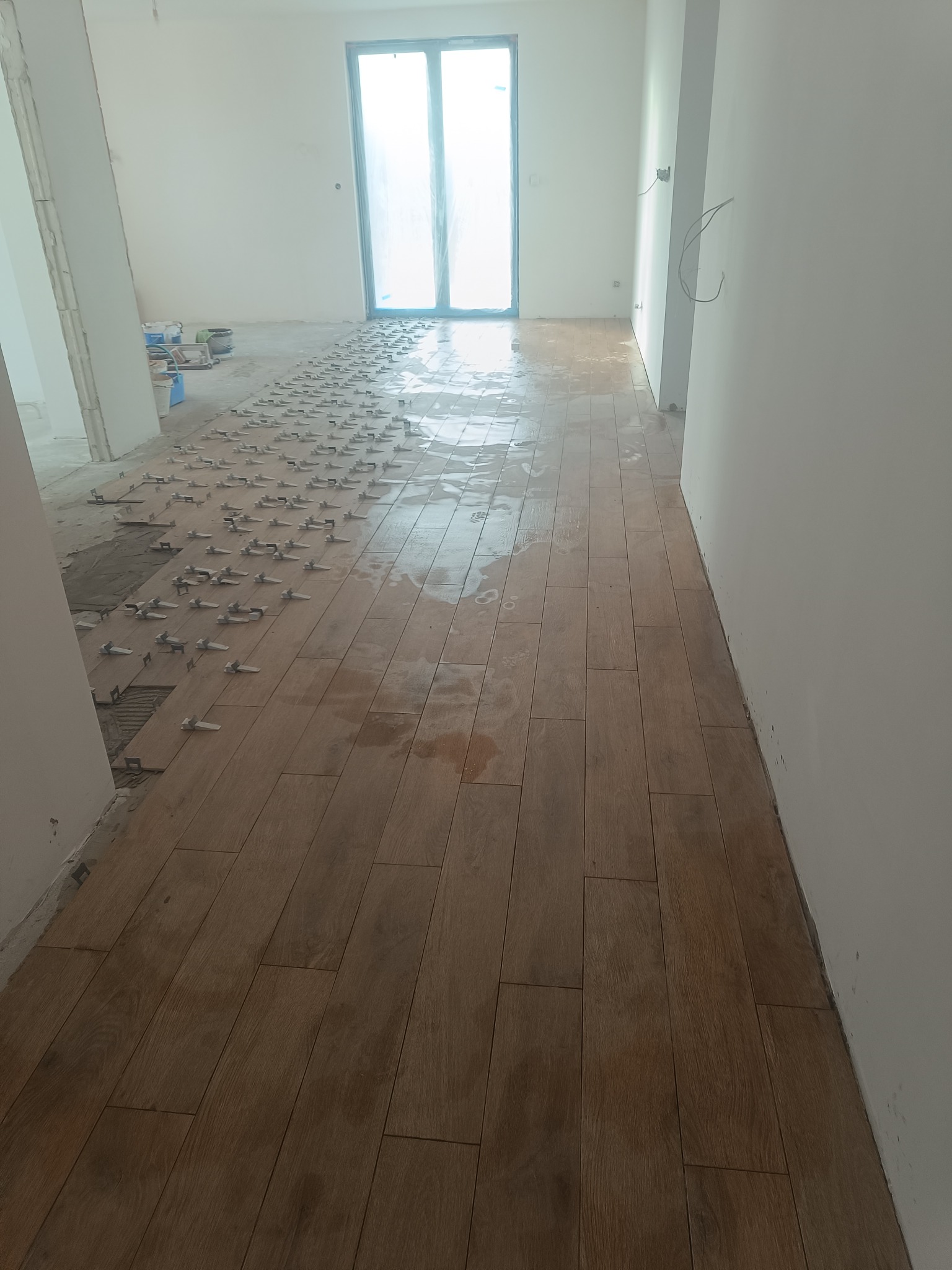 Układanie podłogi z płytek imitujących drewno w jasnym wnętrzu. Widoczne klipsy poziomujące i fragmenty nieukończonej podłogi. W tle okno balkonowe.