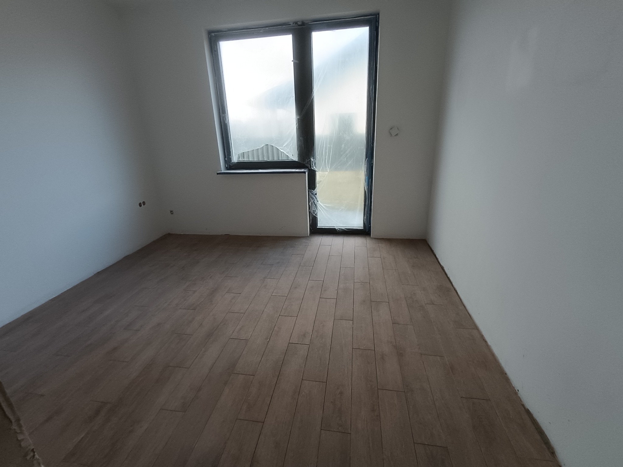 Jasne płytki podłogowe imitujące drewno w pokoju z białymi ścianami i oknem zabezpieczonym folią. Widoczne gniazdka elektryczne.