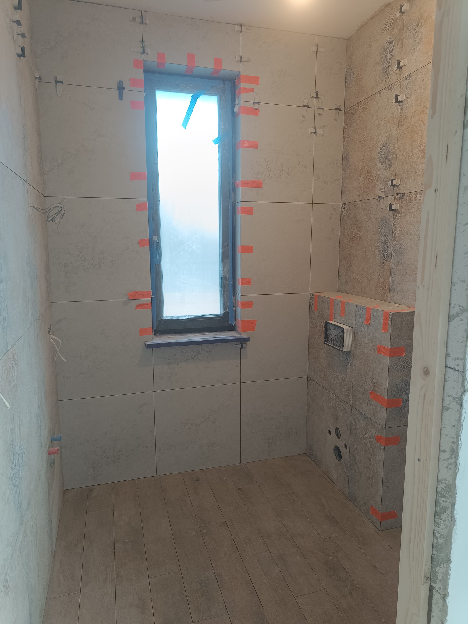 Łazienka w trakcie wykańczania: ułożone płytki ceramiczne na ścianach z oknem, widoczne krzyżyki dystansowe i taśma malarska, drewnopodobna podłoga.