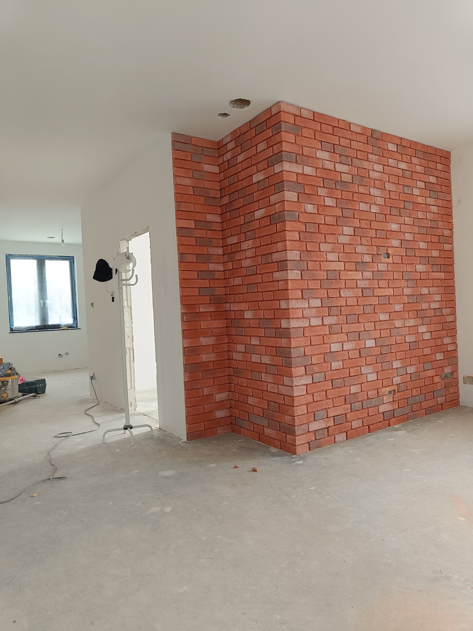 Ściana wykończona czerwoną cegłą klinkierową w rogu pomieszczenia. Widoczne surowe ściany i podłoga, sugerujące etap prac wykończeniowych.