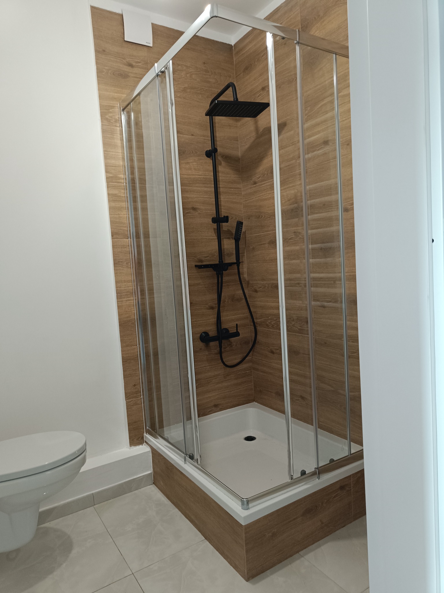 Nowoczesna kabina prysznicowa z czarną armaturą na tle drewnopodobnych płytek w wykończonej łazience. Widoczna toaleta i fragment drzwi.