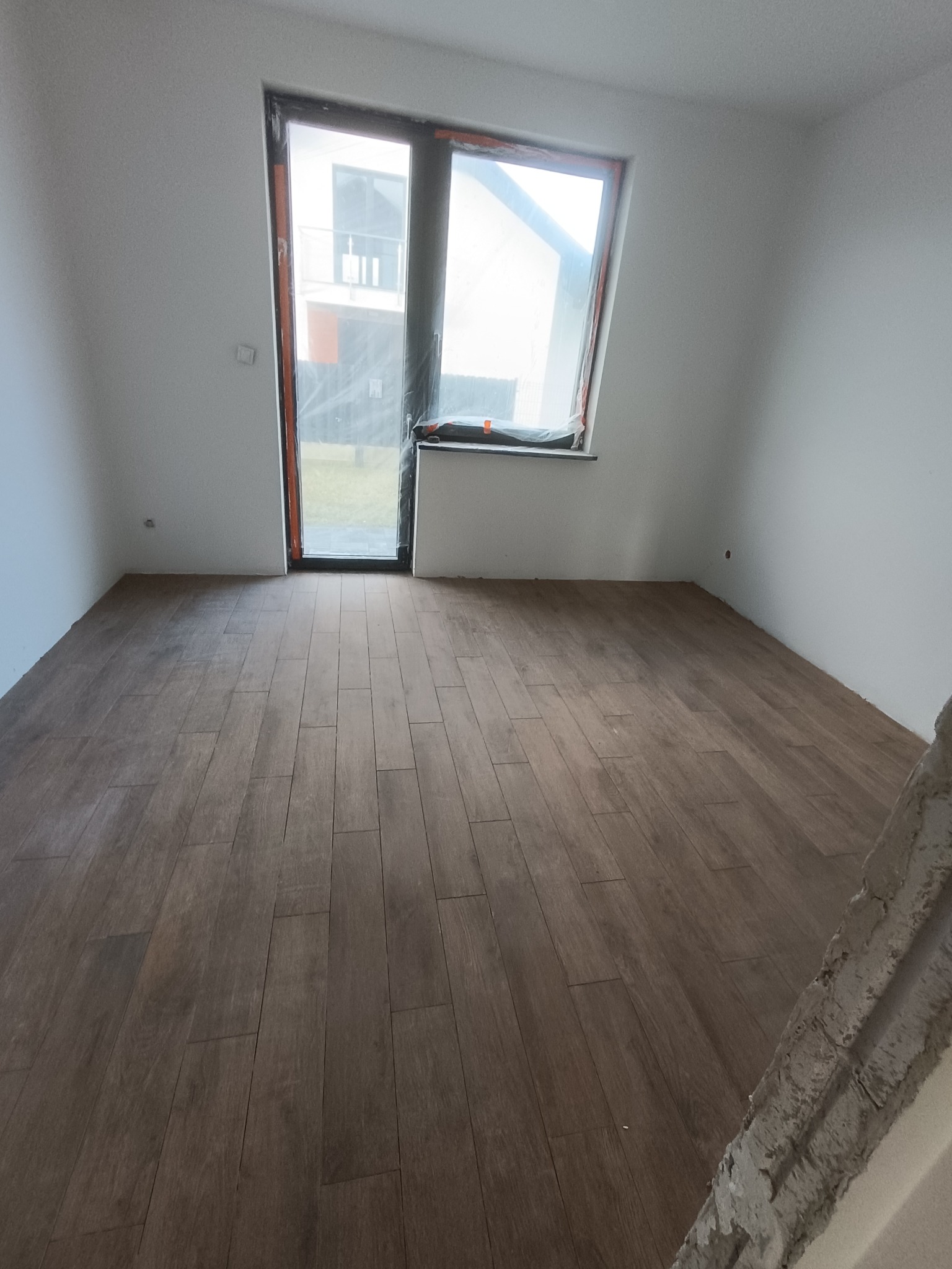 Pokój po remoncie z drewnopodobną podłogą z płytek. Okno zabezpieczone folią. Fragment ściany w prawym dolnym rogu w trakcie prac wykończeniowych.