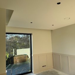 Reshape Mieszko Kurzeja - Jasne wnętrze po malowaniu, z panelami ściennymi w odcieniach beżu i szarości. Widoczne punkty oświetleniowe w suficie i duże okno z widokiem na zieleń.