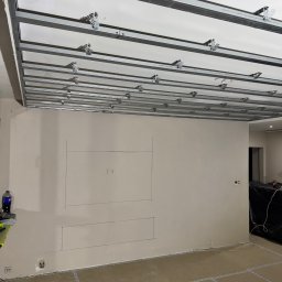 Reshape Mieszko Kurzeja - Montaż stelaża sufitu podwieszanego z płyt karton-gips, widoczne profile i uchwyty. Na ścianie zaznaczone miejsce na TV. Podłoga zabezpieczona taśmą.