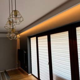 Reshape Mieszko Kurzeja - Jasne wnętrze z drewnianą podłogą i oknami z żaluzjami. Nad oknami subtelne oświetlenie LED. Wisząca lampa z trzema złotymi kloszami dodaje elegancji.