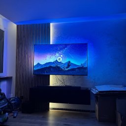 Reshape Mieszko Kurzeja - Nowoczesny salon z podświetlaną ścianą z lameli i telewizorem. Dekoracyjna ściana z efektem betonu. Niebieskie oświetlenie LED tworzy unikalny klimat.