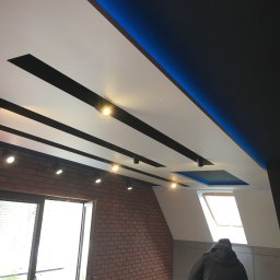 Reshape Mieszko Kurzeja - Nowoczesny sufit podwieszany z oświetleniem LED i reflektorami punktowymi w loftowym wnętrzu z ceglaną ścianą i oknem dachowym. Widoczna osoba w tle.