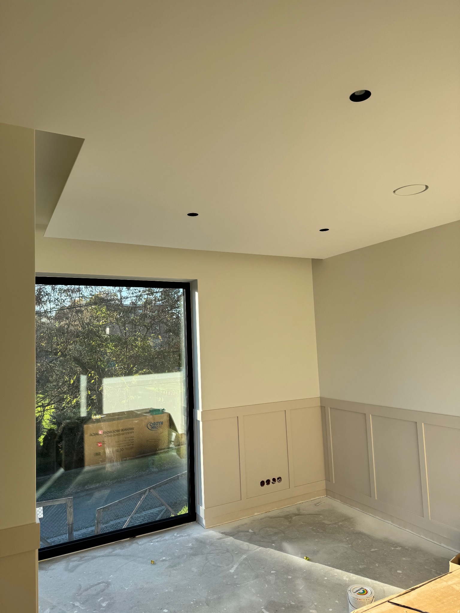 Jasne wnętrze po malowaniu, z panelami ściennymi w odcieniach beżu i szarości. Widoczne punkty oświetleniowe w suficie i duże okno z widokiem na zieleń.