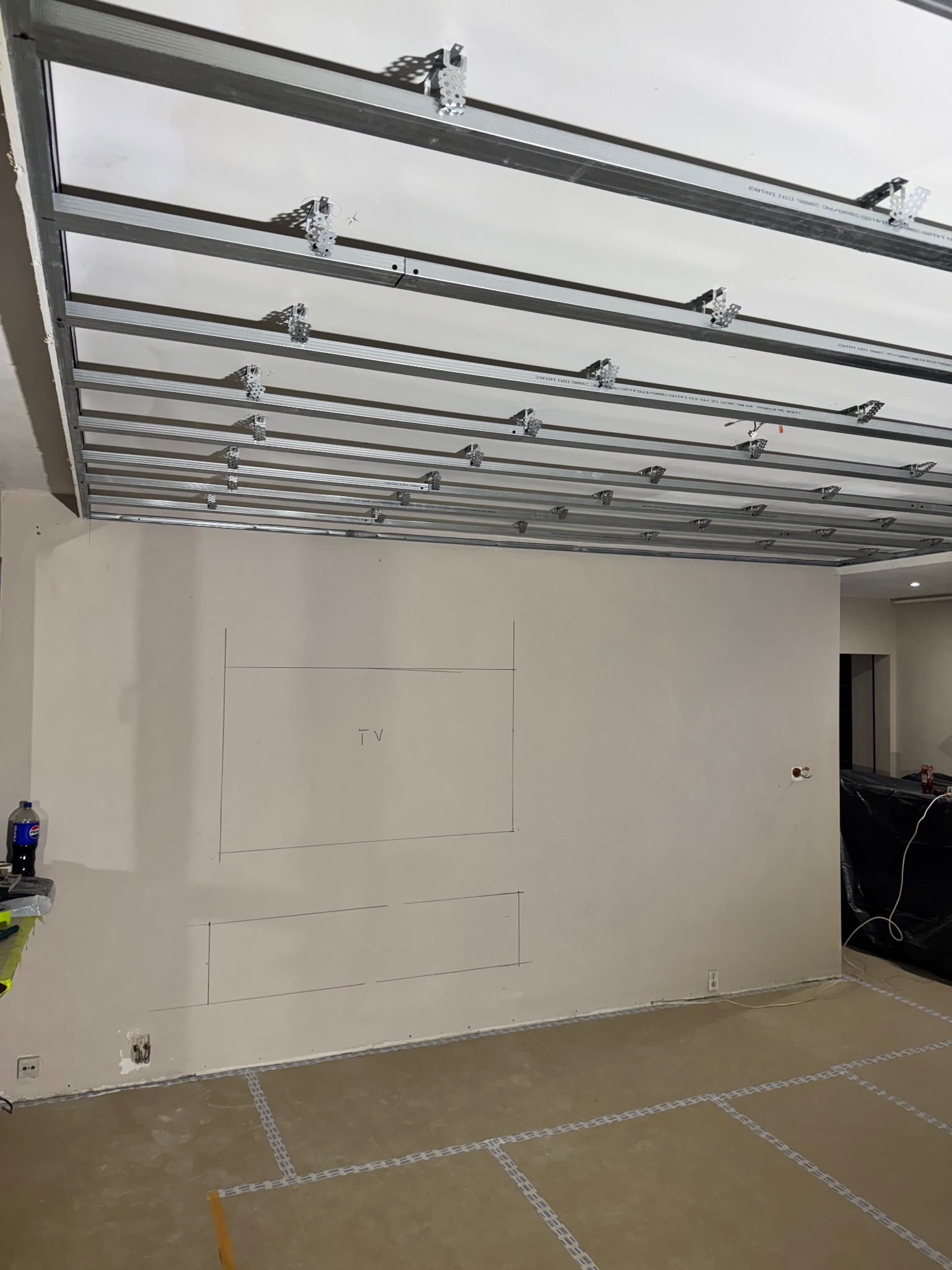 Montaż stelaża sufitu podwieszanego z płyt karton-gips, widoczne profile i uchwyty. Na ścianie zaznaczone miejsce na TV. Podłoga zabezpieczona taśmą.