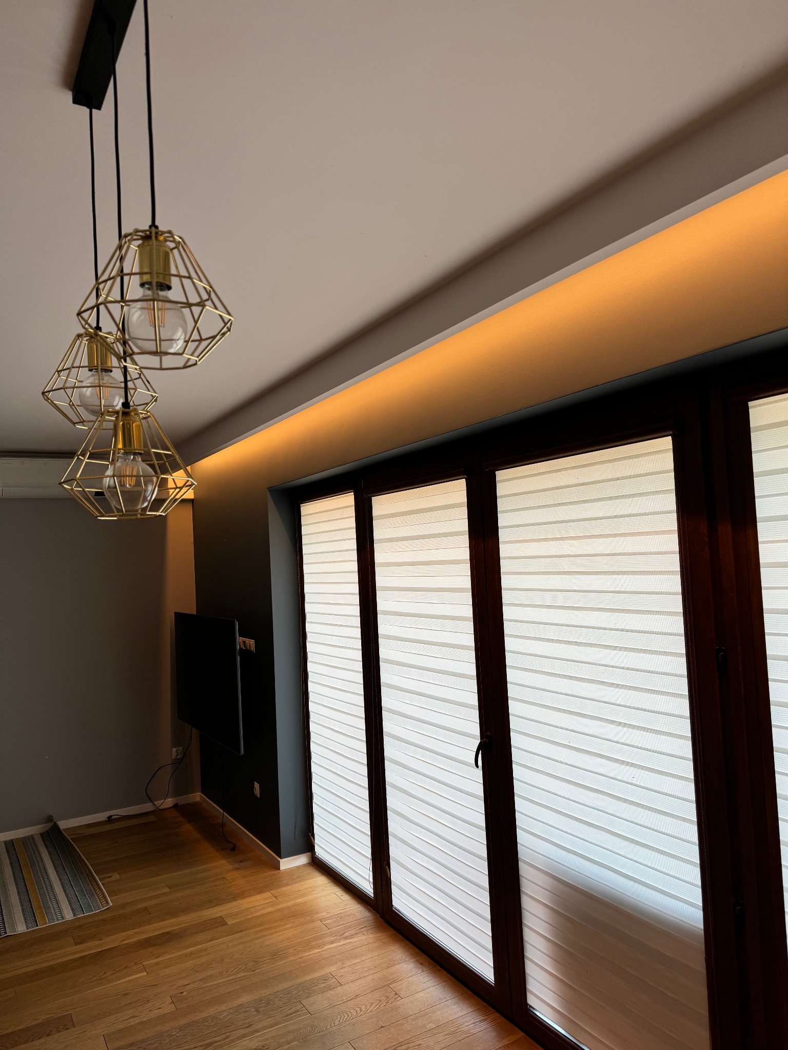 Jasne wnętrze z drewnianą podłogą i oknami z żaluzjami. Nad oknami subtelne oświetlenie LED. Wisząca lampa z trzema złotymi kloszami dodaje elegancji.