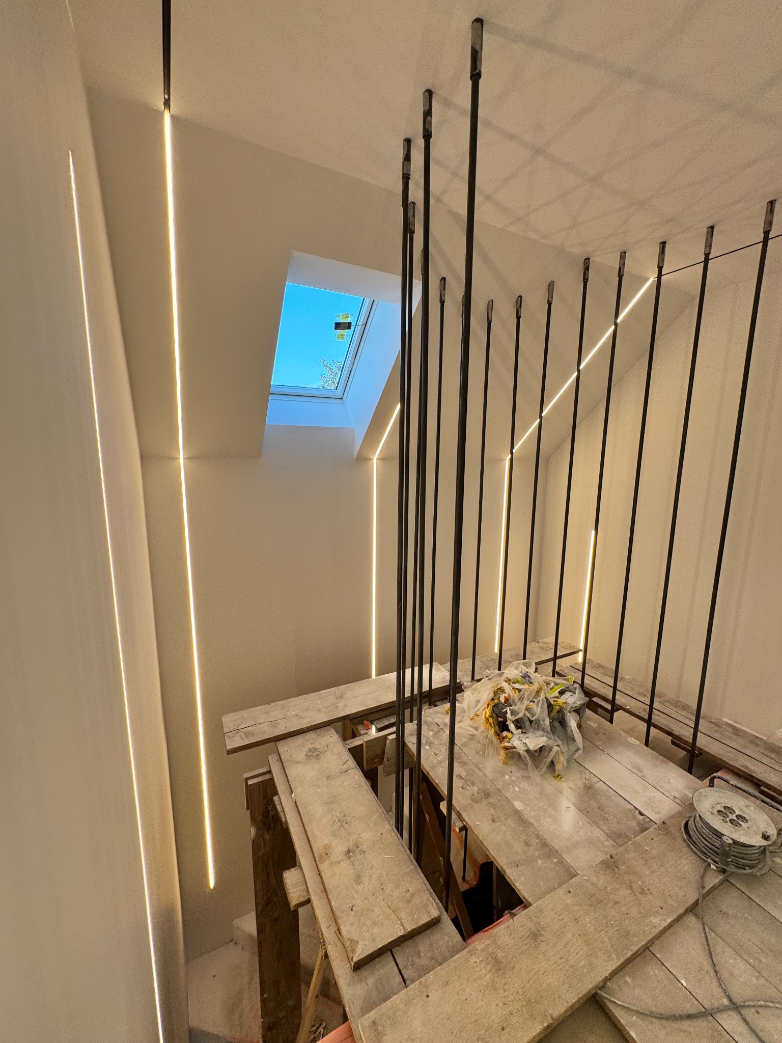 Montaż balustrady schodowej z pionowych prętów i oświetlenia LED wzdłuż ściany, widok na okno dachowe. Prace w toku, widoczne rusztowania i materiały budowlane.