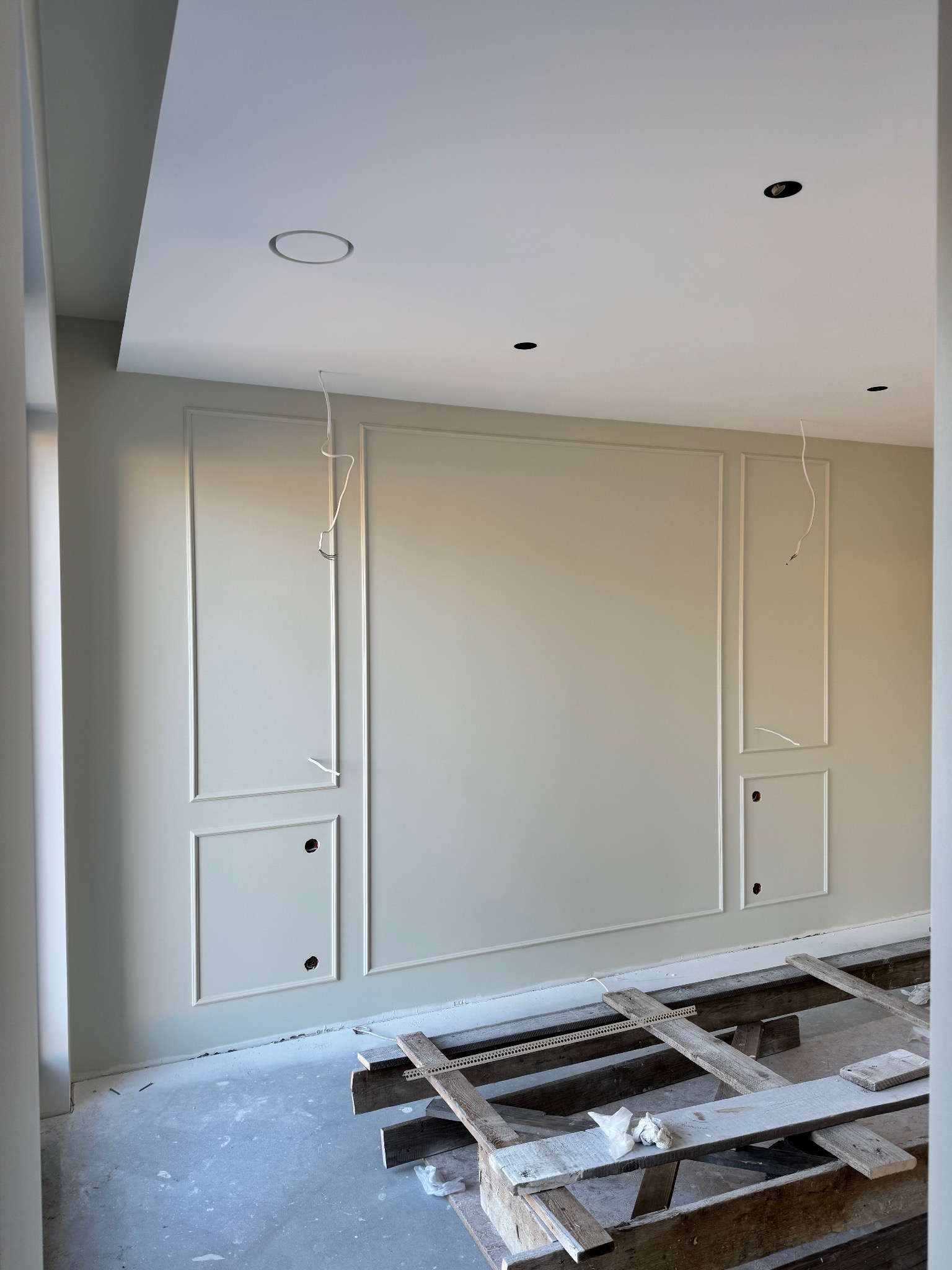 Ściana z dekoracyjnymi panelami pomalowana na jasny kolor, widoczne otwory na gniazdka i wystające przewody. Na podłodze drewniane elementy konstrukcyjne.