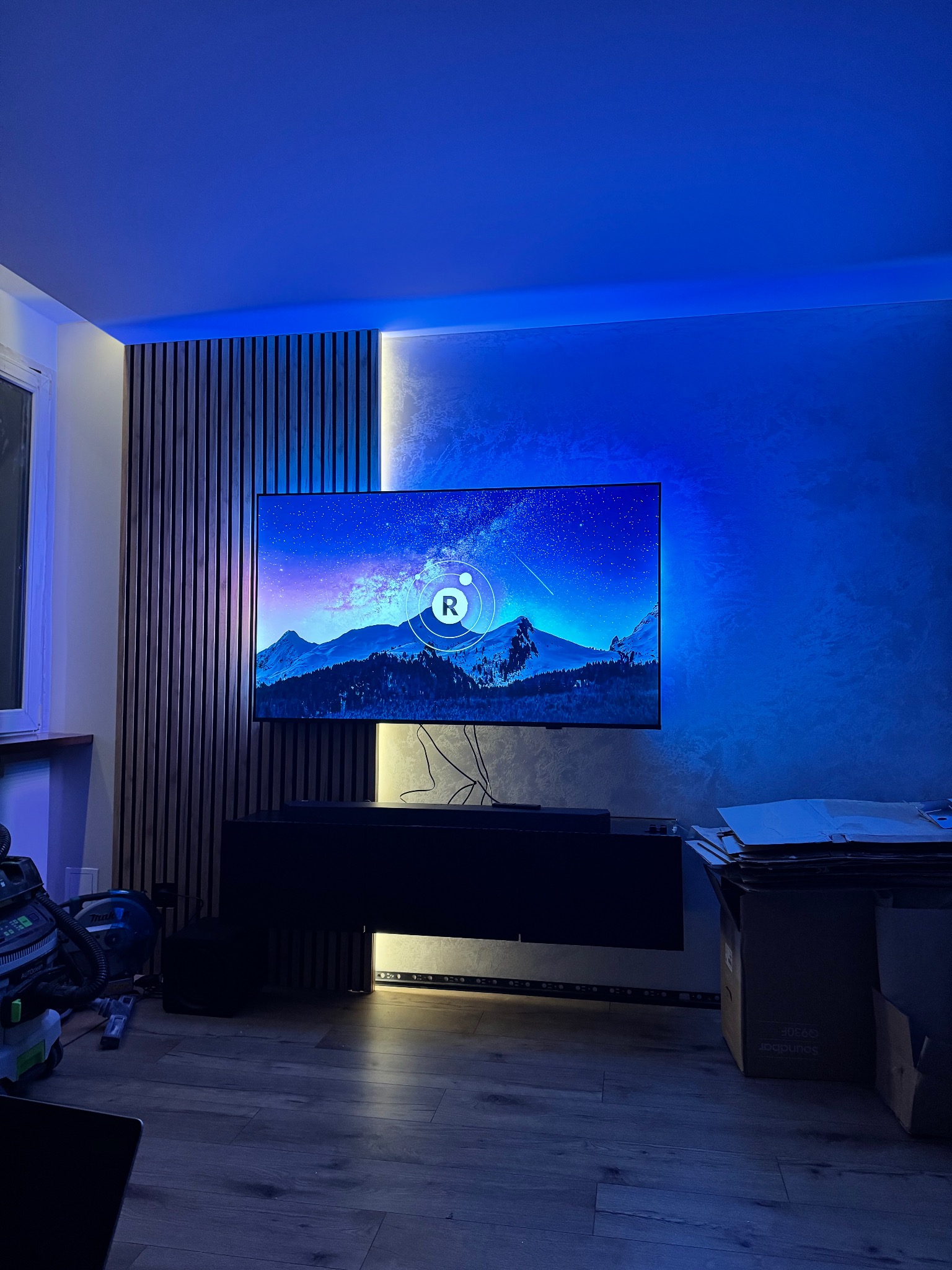 Nowoczesny salon z podświetlaną ścianą z lameli i telewizorem. Dekoracyjna ściana z efektem betonu. Niebieskie oświetlenie LED tworzy unikalny klimat.