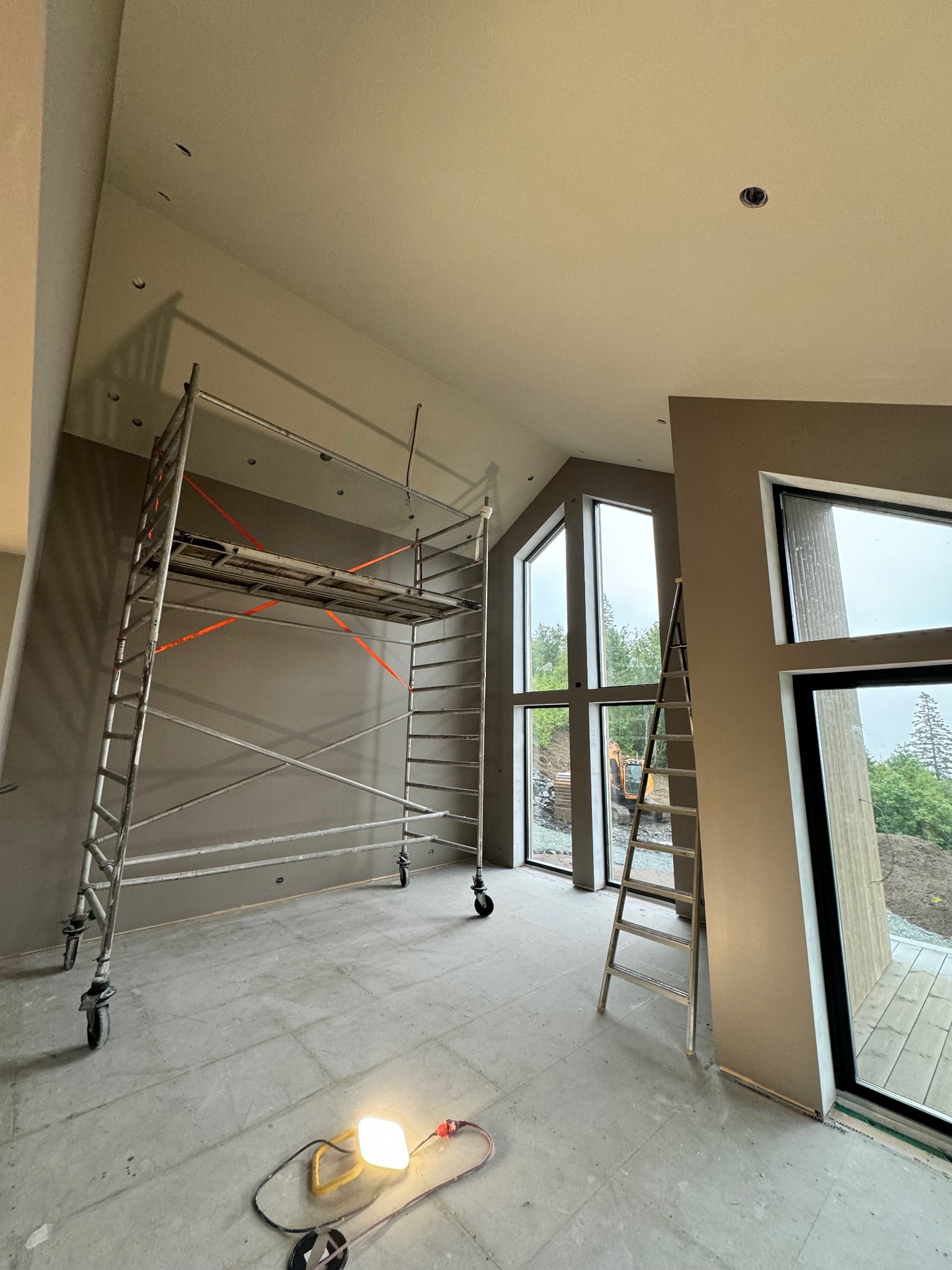 Wnętrze w trakcie remontu: szpachlowana ściana, rusztowanie, drabina oparta o ścianę z oknami. Na podłodze lampa robocza. Widoczne ślady szpachlowania.