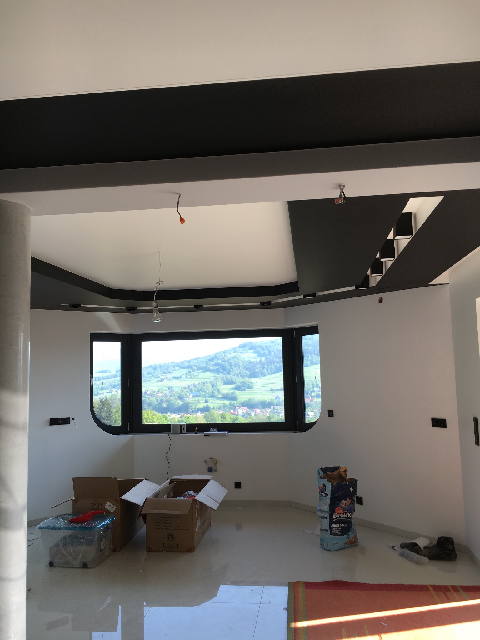 Nowoczesne wykończenie wnętrza z czarnymi elementami sufitu i dużym oknem z widokiem na góry. Widoczne kartony i worek, sugerujące prace wykończeniowe.