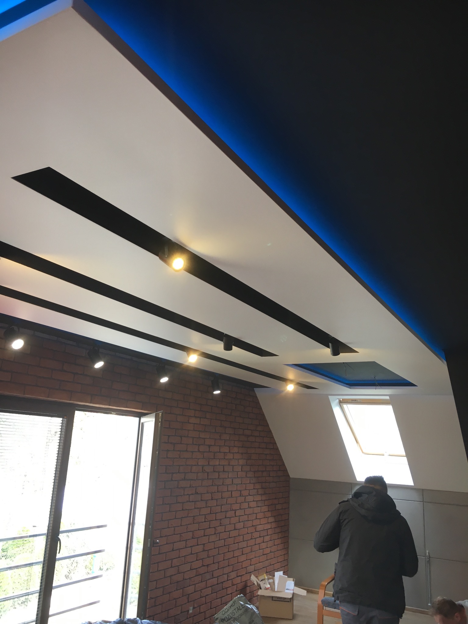 Nowoczesny sufit podwieszany z oświetleniem LED i reflektorami punktowymi w loftowym wnętrzu z ceglaną ścianą i oknem dachowym. Widoczna osoba w tle.