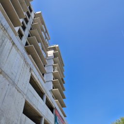 HUBBUD - Surowy budynek żelbetowy w trakcie budowy, widok z dołu na szkielet konstrukcji i błękitne niebo. Na budynku widoczny baner reklamowy.