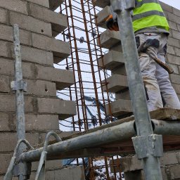 HUBBUD - Praca murarska: robotnik w kasku i kamizelce na rusztowaniu, w trakcie wznoszenia ściany z bloczków betonowych z widocznym zbrojeniem. Zima, śnieg w tle.