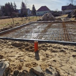 HUBBUD - Zbrojenie fundamentów domu jednorodzinnego, widok na siatkę zbrojeniową, szalunek i teren budowy w słoneczny dzień. Pomarańczowa rura pionowo w ziemi.