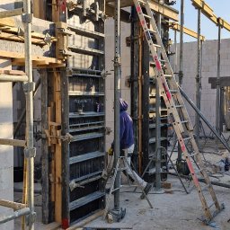 HUBBUD - Szare szalunki pionowe z drewnianymi wzmocnieniami, drabina oparta o konstrukcję, pracownik w tle, widok na plac budowy w słoneczny dzień.