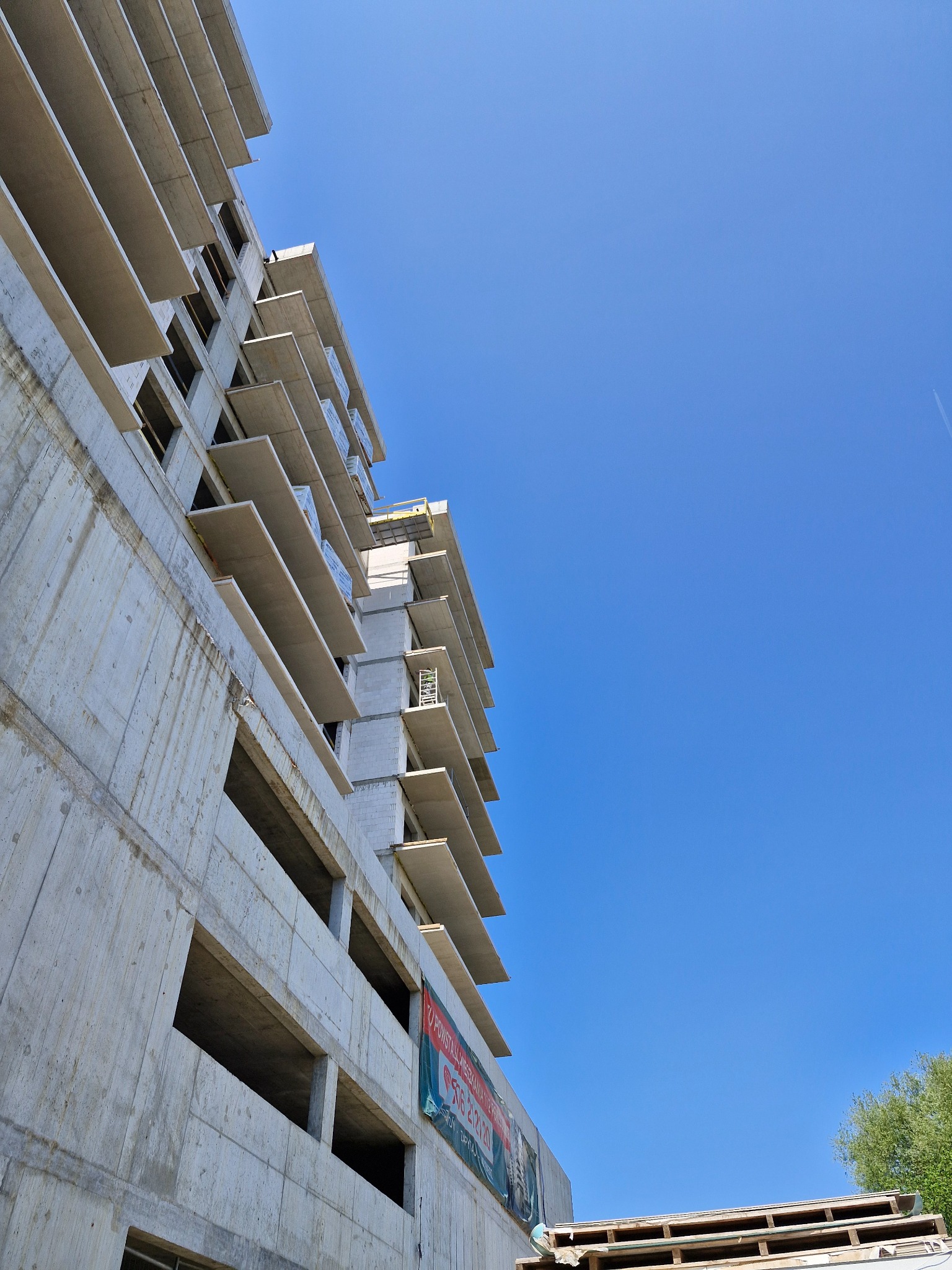 Surowy budynek żelbetowy w trakcie budowy, widok z dołu na szkielet konstrukcji i błękitne niebo. Na budynku widoczny baner reklamowy.