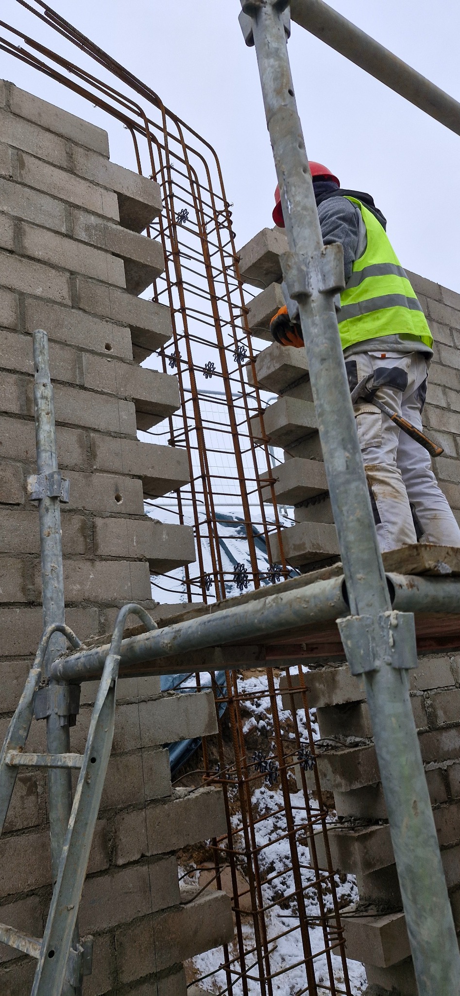 Praca murarska: robotnik w kasku i kamizelce na rusztowaniu, w trakcie wznoszenia ściany z bloczków betonowych z widocznym zbrojeniem. Zima, śnieg w tle.