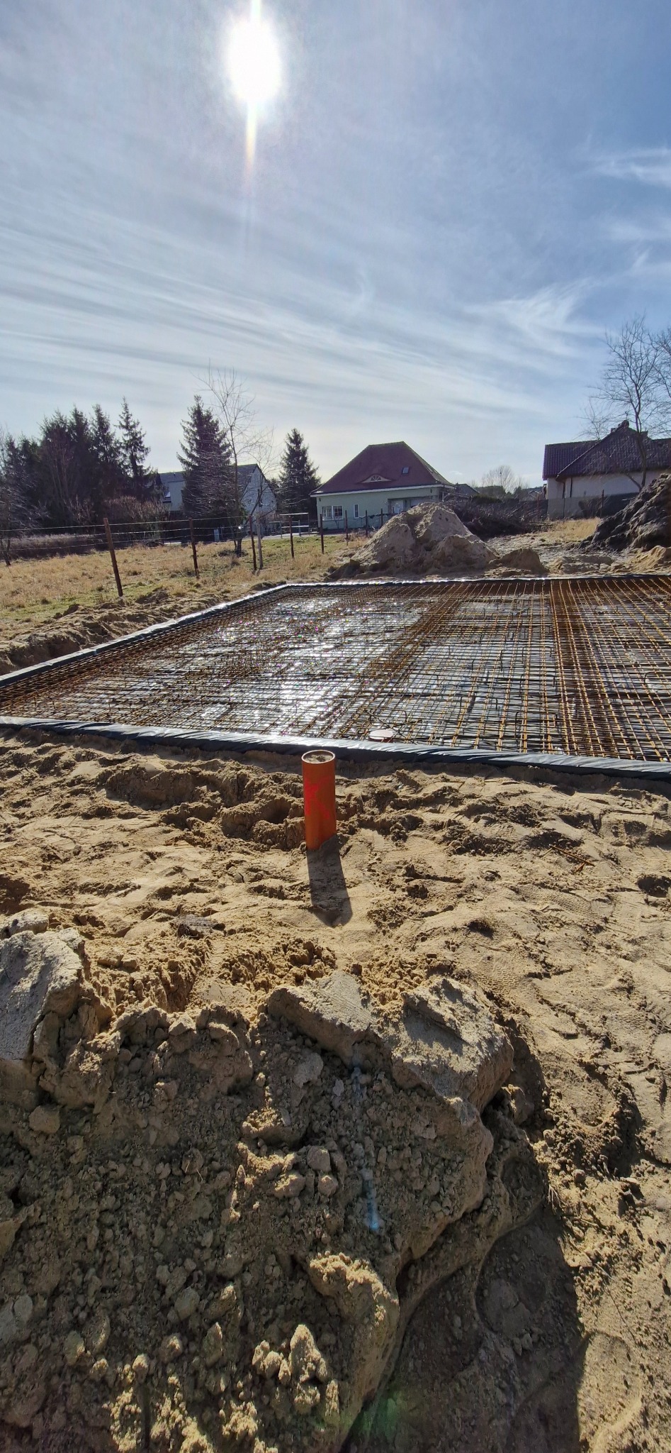 Zbrojenie fundamentów domu jednorodzinnego, widok na siatkę zbrojeniową, szalunek i teren budowy w słoneczny dzień. Pomarańczowa rura pionowo w ziemi.