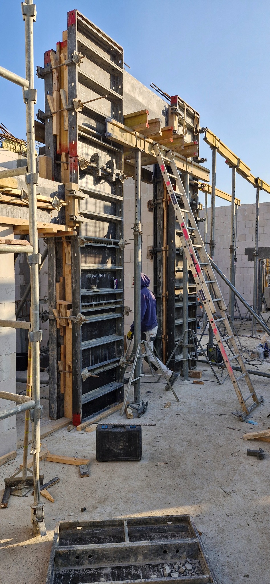 Szare szalunki pionowe z drewnianymi wzmocnieniami, drabina oparta o konstrukcję, pracownik w tle, widok na plac budowy w słoneczny dzień.