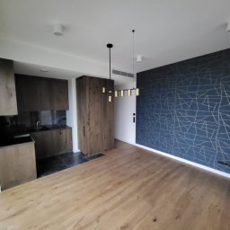 Mateusz Marcinkiewicz - Nowoczesne wnętrze po remoncie: drewniana podłoga, zabudowa kuchenna z ciemnego drewna i ściana z geometrycznym wzorem w odcieniach granatu i złota.