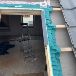 Mateusz Marcinkiewicz - Montaż okna dachowego z uszczelnieniem i folią paroizolacyjną. Widoczna konstrukcja dachu i drabina w surowym wnętrzu poddasza.